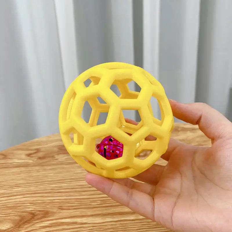 A hand holds a yellow natural rubber dog chew ball with a pink bell, shaped like a honeycomb.