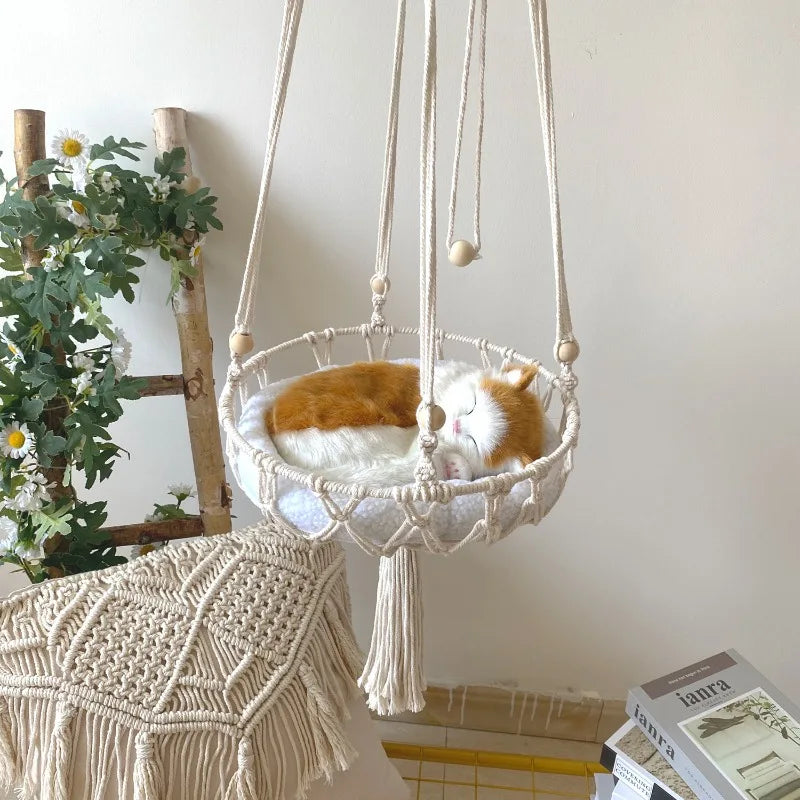 A hand-woven cotton cat hammock with a white cushion is suspended by macrame ropes.