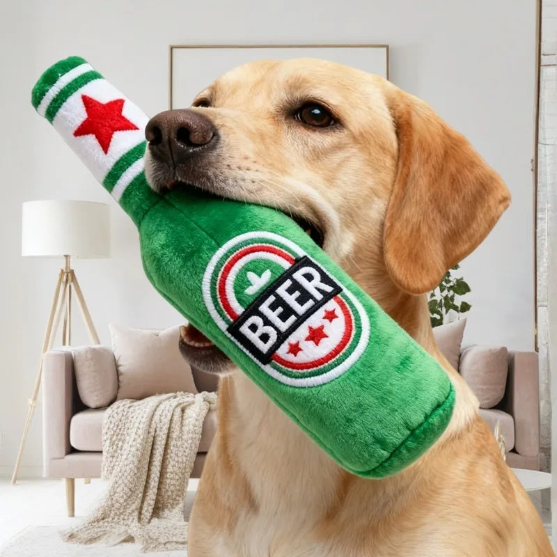 A golden retriever holds a green squeaky dog toy shaped like a beer bottle with red stars and black lettering.
