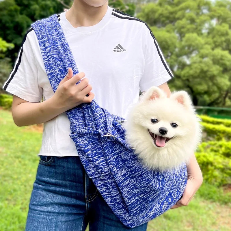 A person is holding a blue and white patterned fabric sling carrier for a small white dog outdoors.