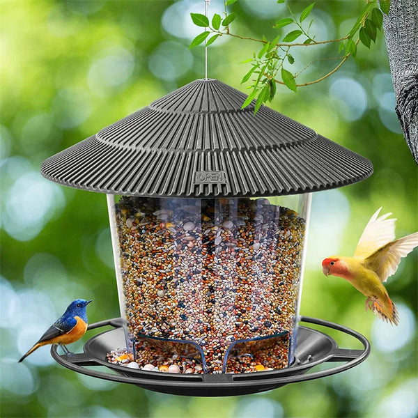 A hanging bird feeder with a conical roof filled with seeds, suspended from a branch against a blurred green background.
