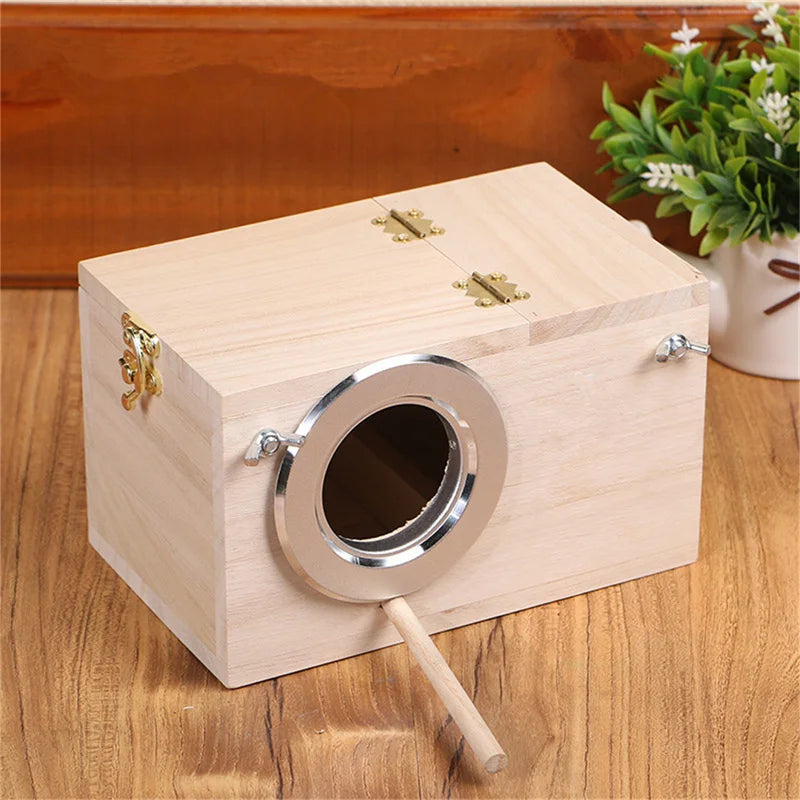 A light wood bird nest box with a circular entrance and brass hinges sits on a wooden surface.