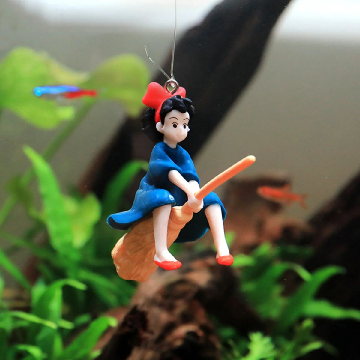 A small figurine of a mermaid with red hair and blue skin is suspended by a transparent line, set against a blurred green bac