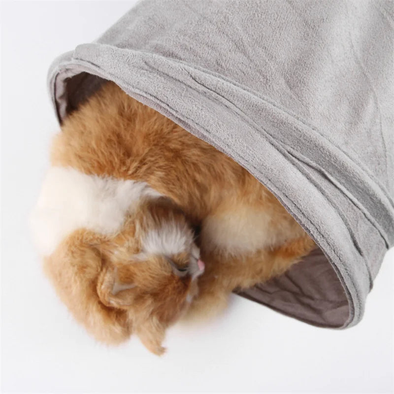 A fluffy orange and white kitten is peeking out of a gray foldable S-shape cat tunnel.
