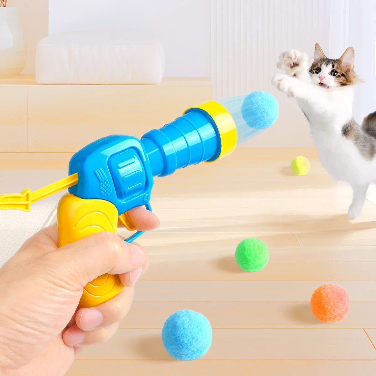 A hand holds a blue and yellow cat toy ball launcher, which is firing a blue plush ball toward a cat.