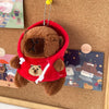 Braune Capybara-Plüschpuppe mit rotem Hoodie und weißem Zierrand, an einer goldenen Kette aufgehängt.