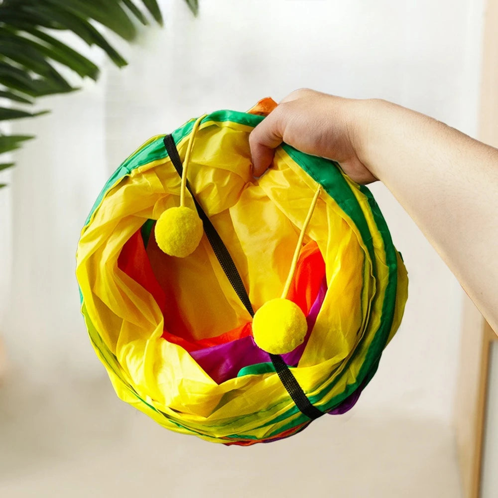 A hand is holding a bright yellow rainbow cat tunnel with multiple entrances, featuring colorful fabric and yellow pom-poms.