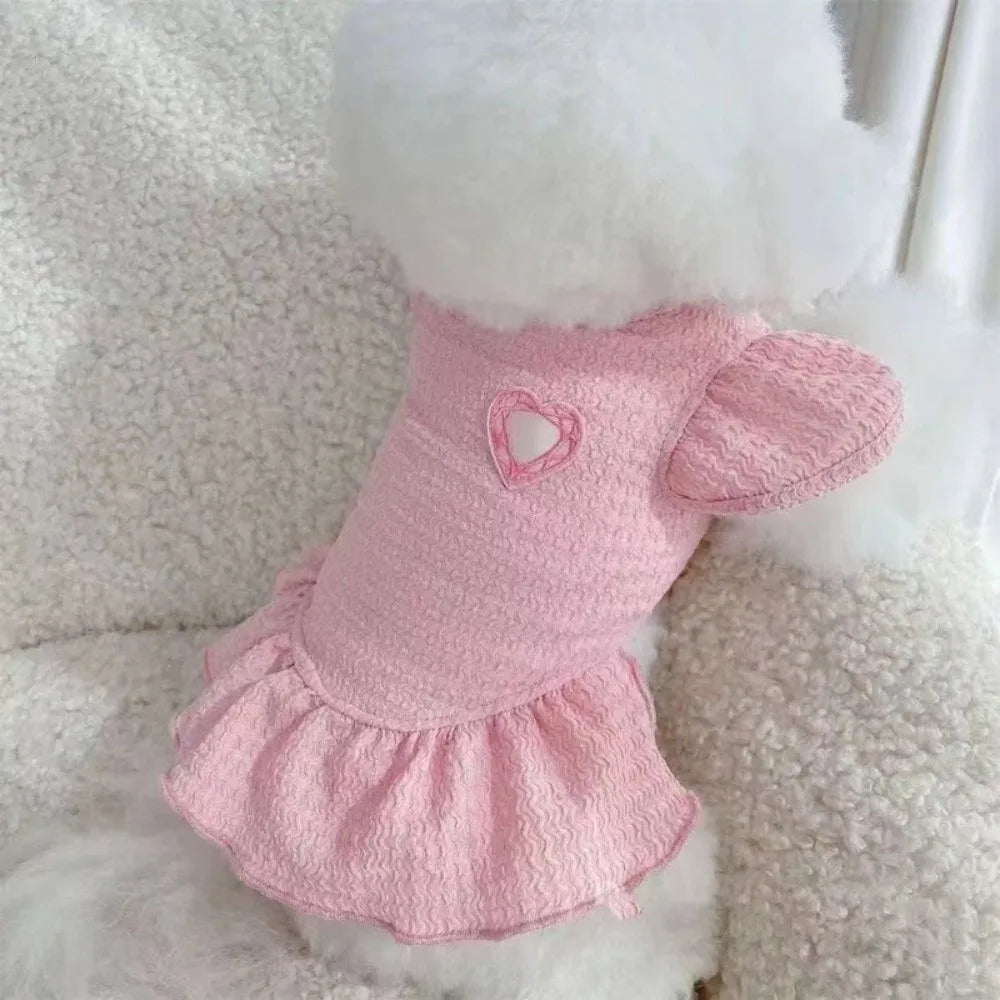 A pink pet dress for a dog featuring flutter sleeves and a heart detail, displayed against a white fluffy background.