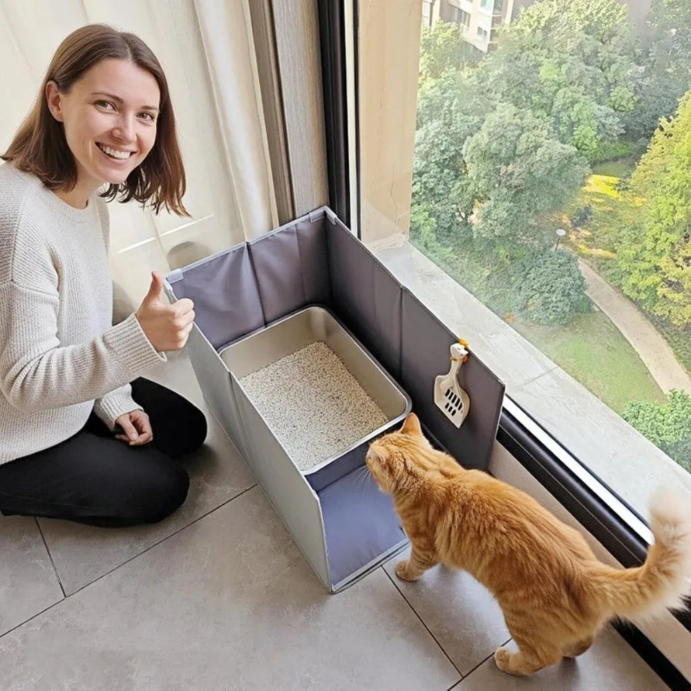 A woman sits next to a foldable cat litter box enclosure with a splash guard, while a orange cat looks out of the box.