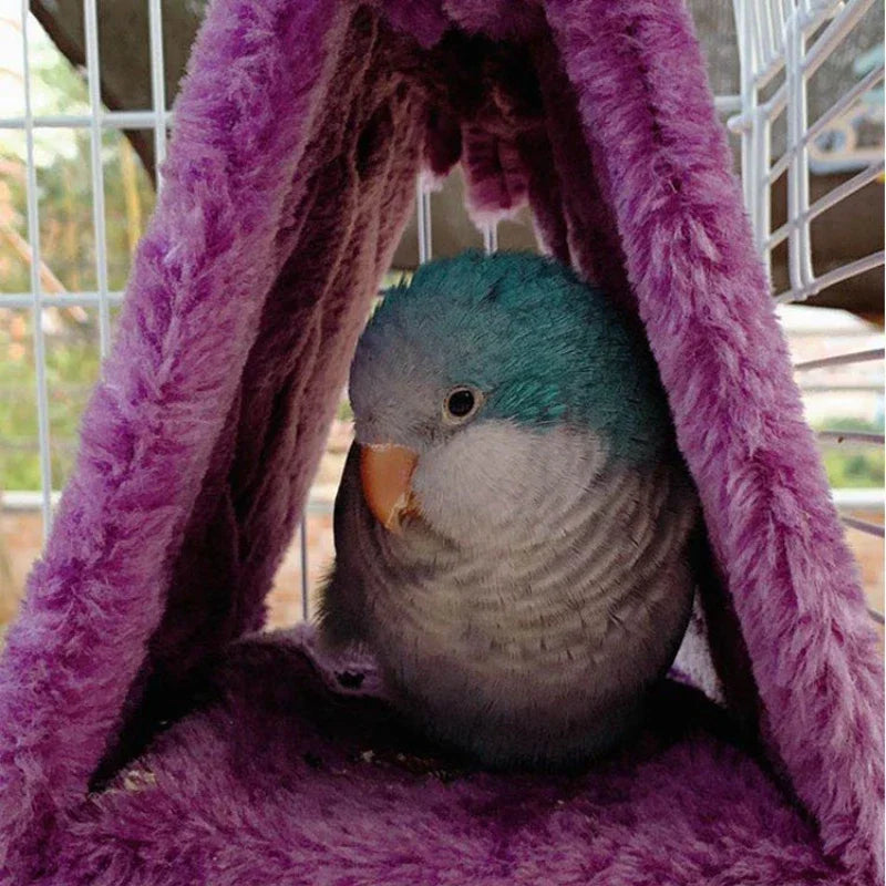 A blue and gray budgerigar sits inside a purple plush hammock nest in a wire cage.