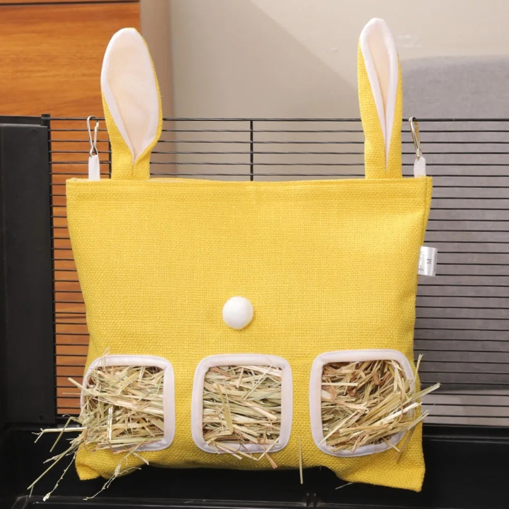 A bright yellow multi-hole hay feeder bag for small animals with white ear flaps and straw bedding.
