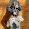 A person is holding a beige portable pet carrier sling bag with a brown strap and a small white fluffy dog.
