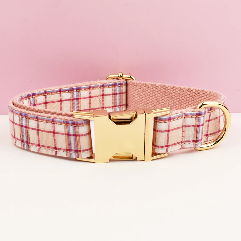 A pink plaid dog collar with a gold buckle and leather strap is displayed against a light pink background.