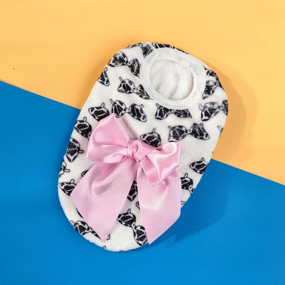 A white knit pet sweater with black dog faces and a pink satin bow, displayed on a yellow and blue background.