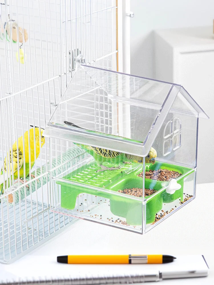 A clear plastic parrot feeder and bird bath accessory is placed in front of a wire cage.