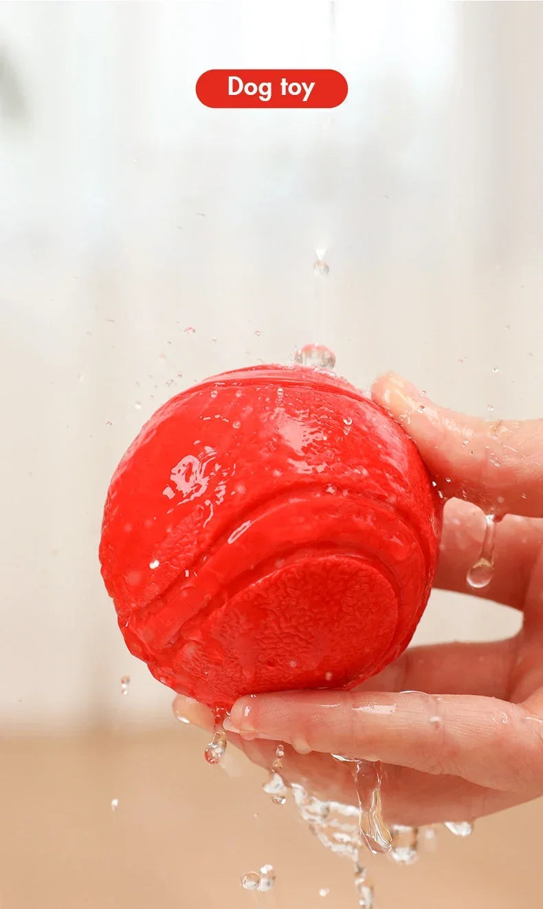 A hand is holding a bright red rubber dog toy ball, with water droplets splashing around it.
