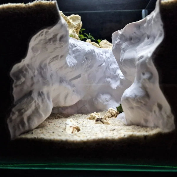 A white 3D rock cave aquarium ornament with natural terrain design set in sand.