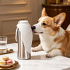 A light brown and white dog is looking at a stainless steel portable water bottle with a built-in bowl on a marble surface.
