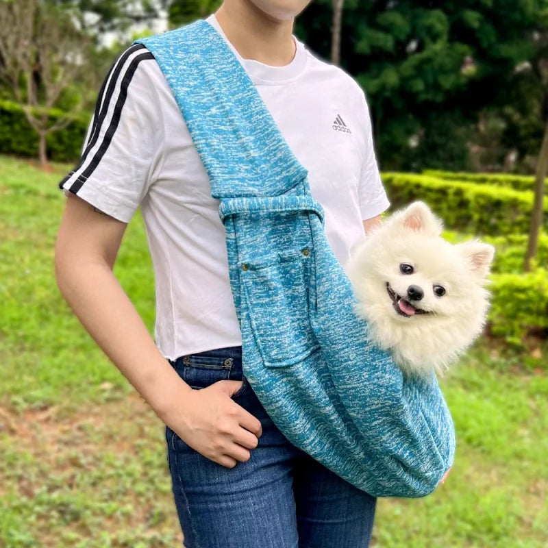 A person wearing a white t-shirt and jeans is holding a blue patterned pet sling carrier for a small dog.