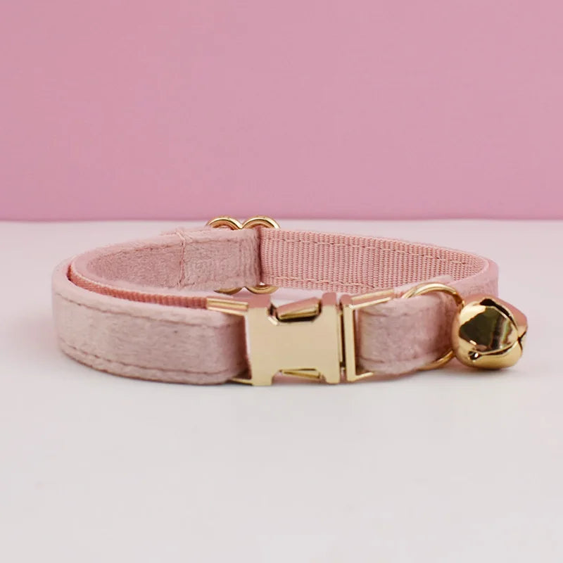 A pink velvet cat collar with a gold-toned buckle and bell resting on a white surface.