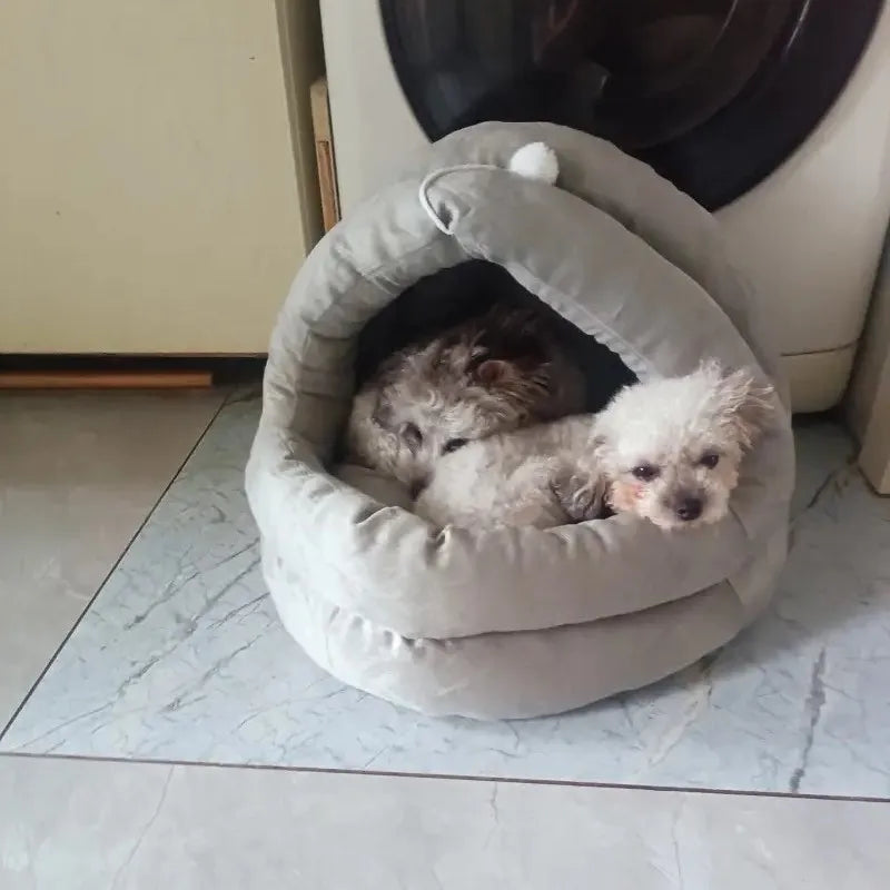 A fluffy gray cat is resting inside a soft, oval-shaped plush nest bed with a hood.