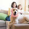 A light-colored dog is lying on a couch with two people smiling behind it, wearing a pink ear muff hat.
