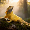 A bearded dragon wearing a yellow hooded costume perched on a mossy log in a natural, misty setting.