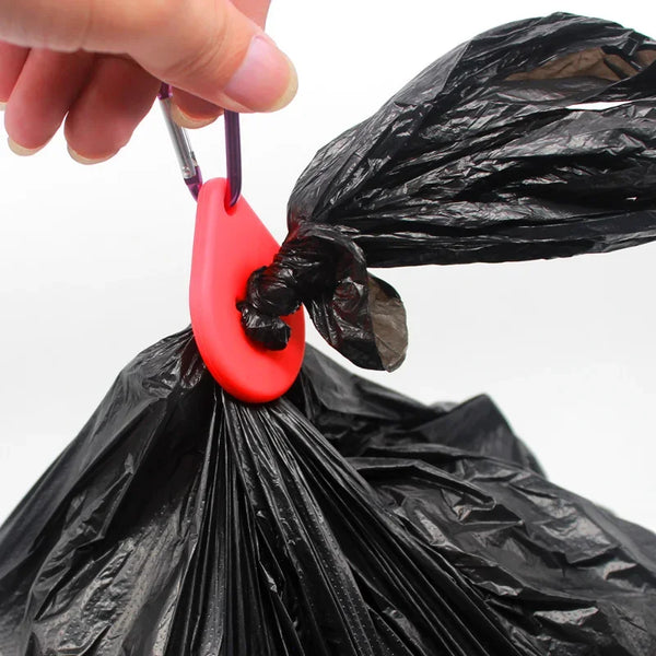 A hand is holding a red clip attached to a black plastic bag, which is a hands-free dog poop bag dispenser.