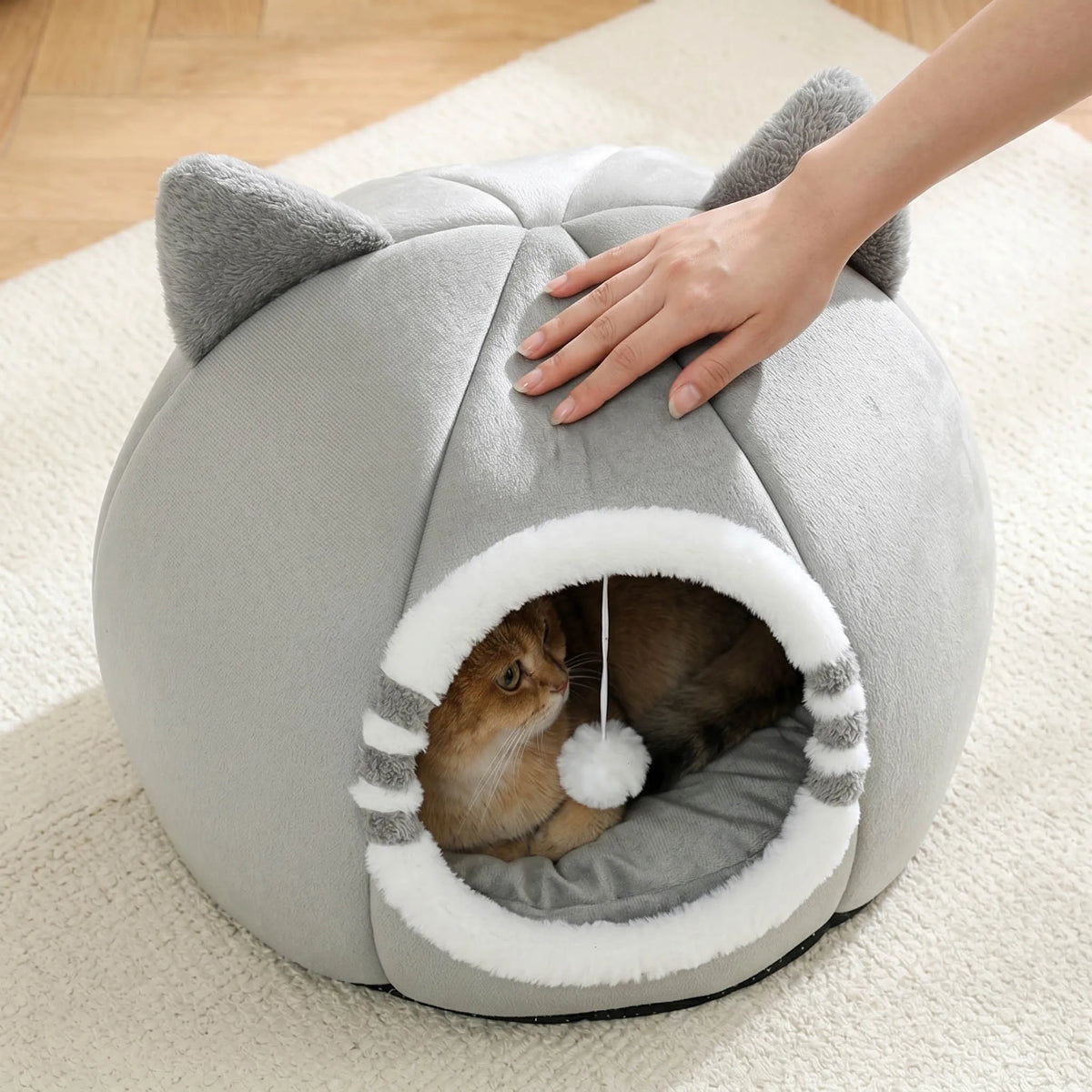 A gray plush cartoon cat cave bed with an entrance, being touched by a person's hand.