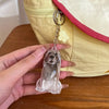 A clear glass keychain shaped like a dog is held against a yellow fabric pouch with a pink trim.