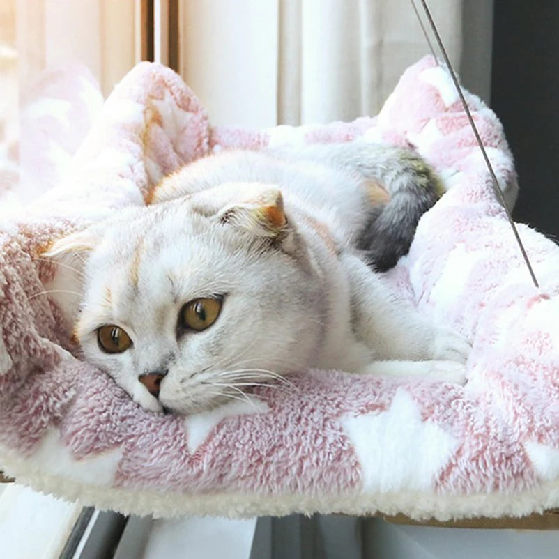Un chat gris et blanc duveteux se détend dans un lit-hamac texturé rose fixé à la fenêtre.