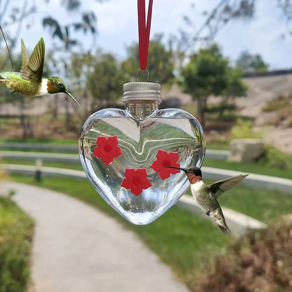 A clear heart-shaped glass feeder hangs, with a hummingbird visible near the red flower decorations.