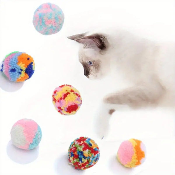 A fluffy gray cat interacts with six colorful plush wool balls of various sizes and colors.
