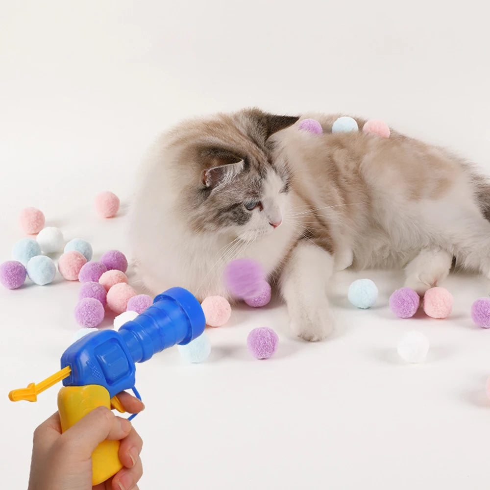 A hand is holding a blue interactive cat toy gun that is spraying soft pink and white plush balls onto a cat.