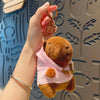 Eine Hand hält eine braune Capybara-Plüschpuppe mit pinkem Hoodie und goldenen Flügeln vor einem hellblauen gemusterten Hinte