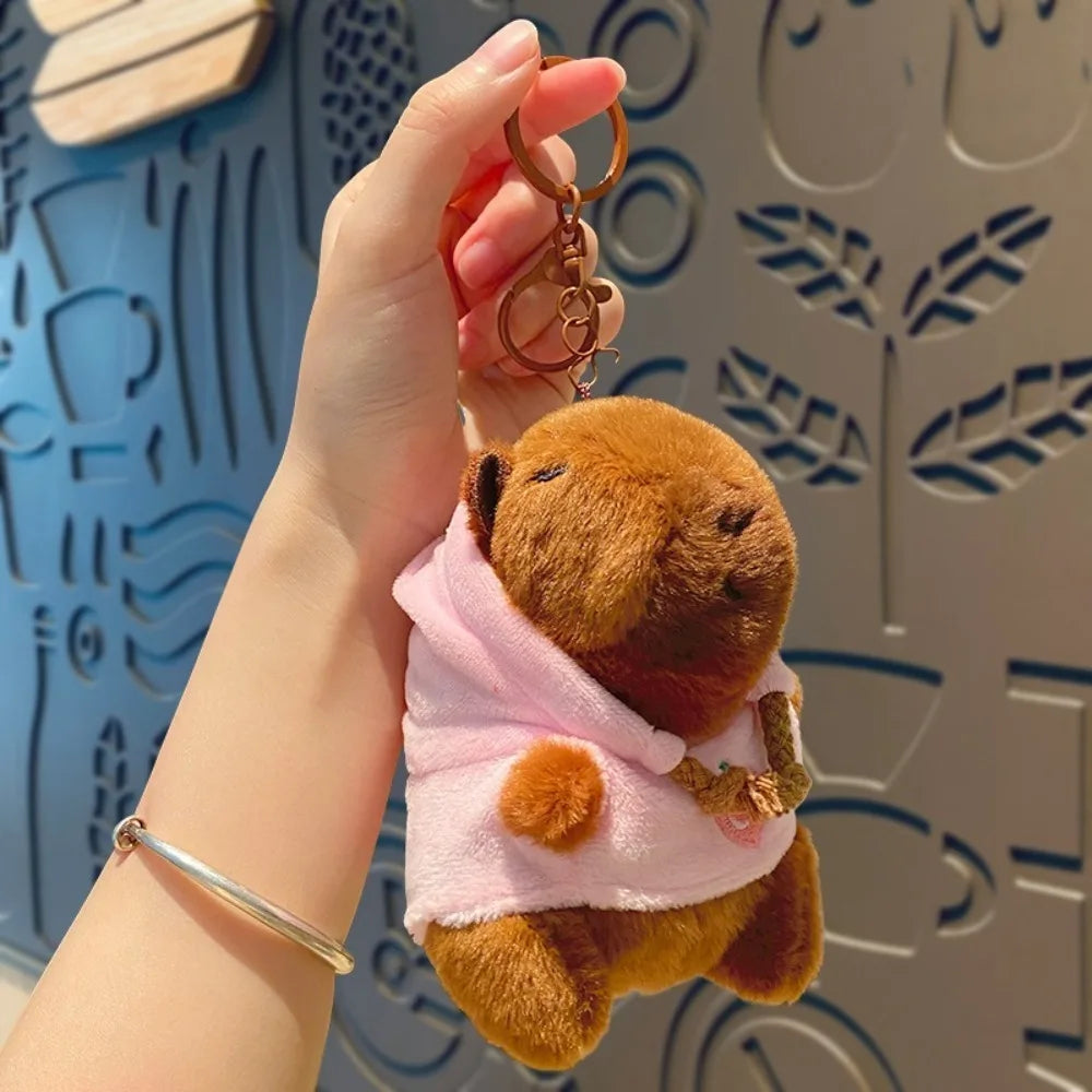 Eine Hand hält eine braune Capybara-Plüschpuppe mit pinkem Hoodie und goldenen Flügeln vor einem hellblauen gemusterten Hinte