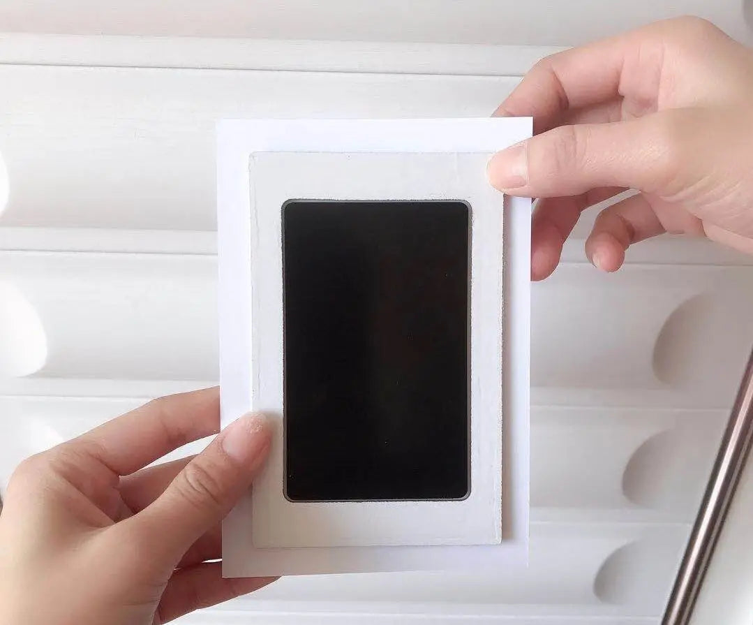 A hand is holding a white frame containing a black print, likely for pet paw print and handprint supplies.