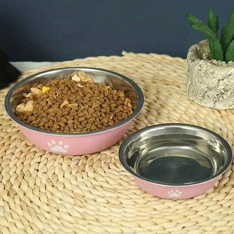 A pink stainless steel pet bowl with a non-slip silicone base filled with dry pet food on a woven mat.