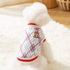A white fluffy dog toy wearing a red and white plaid knit sweater with a bear design.
