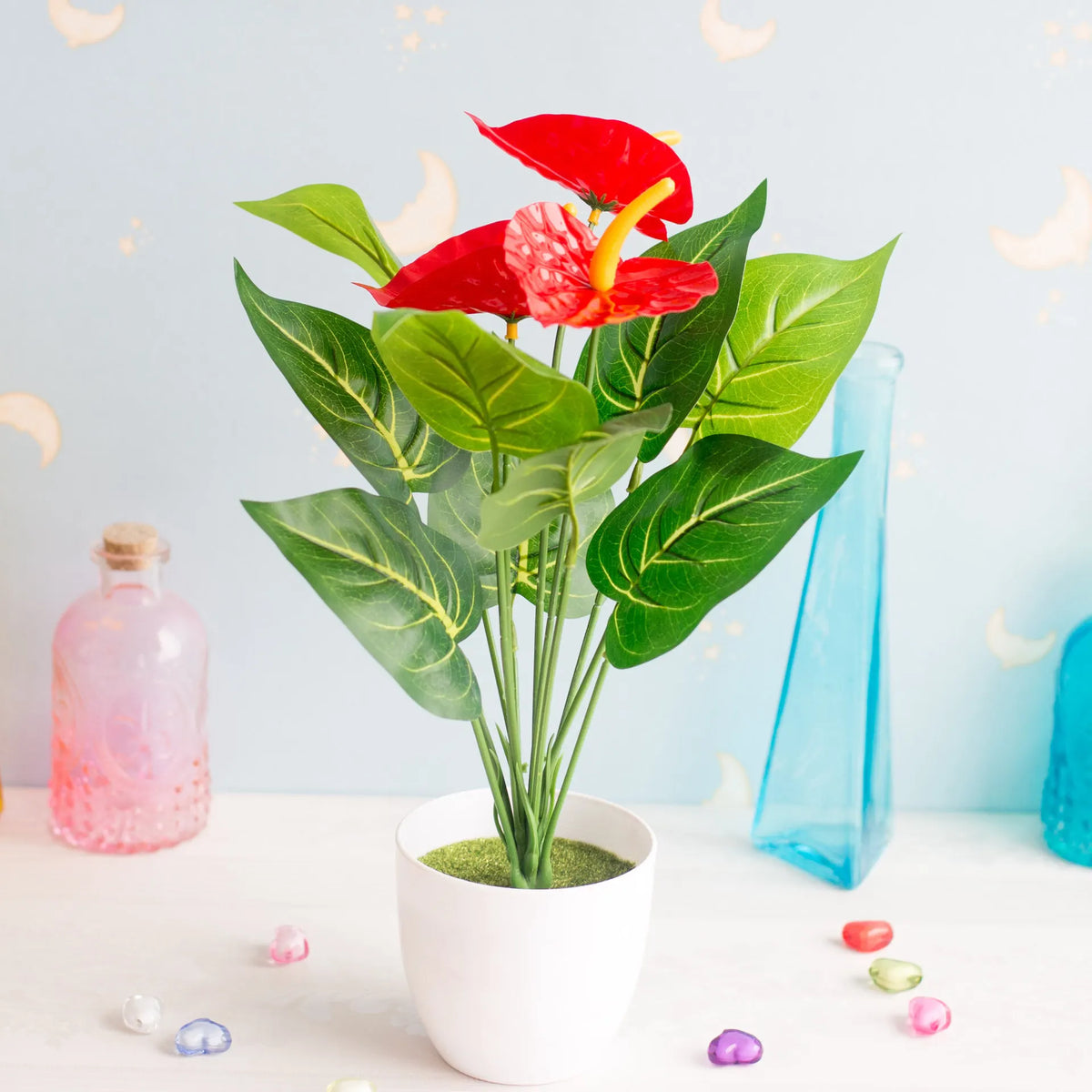 A vibrant red artificial anthurium plant in a white pot set against a light blue background.