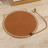 A round, brown cat scratching mat with a rope handle and self-play ball resting on a light wood floor.