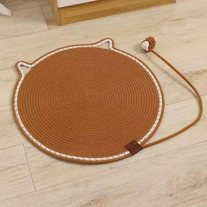 A round, brown cat scratching mat with a rope handle and self-play ball resting on a light wood floor.