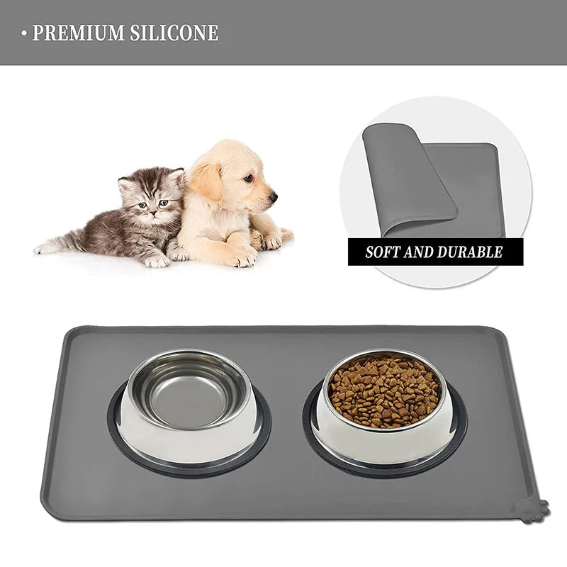 A gray silicone pet feeding mat with raised edges, shown next to two small puppies and a bowl of dry food.