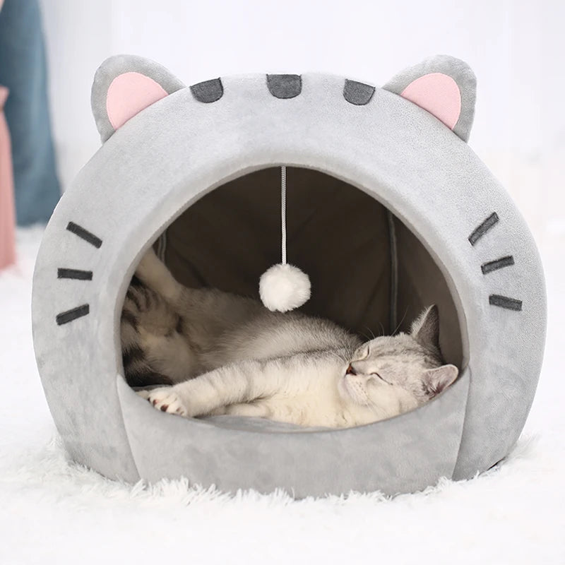 Un lit pour chat gris en forme de grotte avec des touffes d'oreilles et des marques faciales, contenant un chaton blanc endor