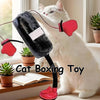 Eine weiße Katze spielt mit einem rosa und einem schwarzen Box-Spielzeug in einem hellen Raum.