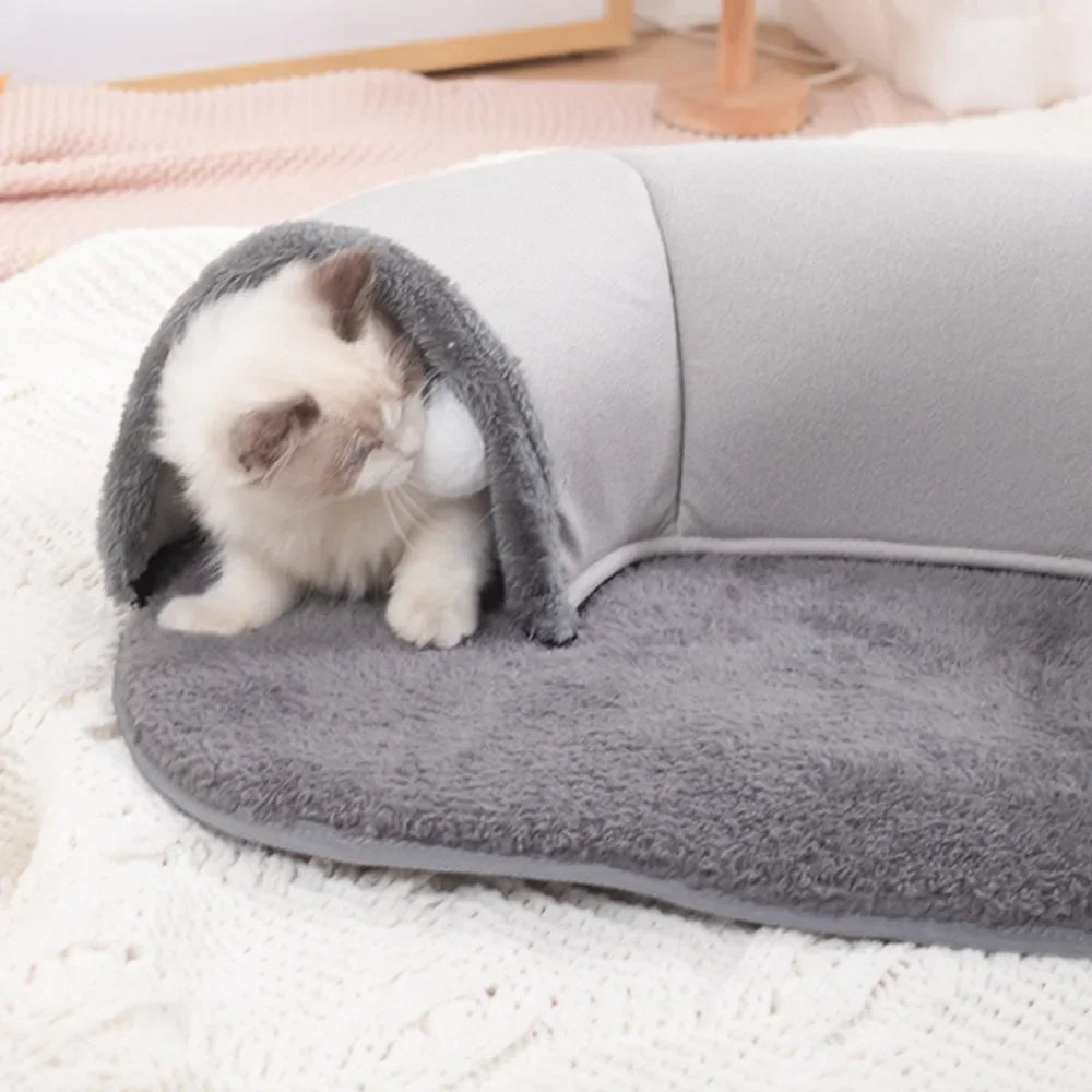 A small white kitten is nestled inside a gray oval cat tunnel and cozy bed nest on a white blanket.