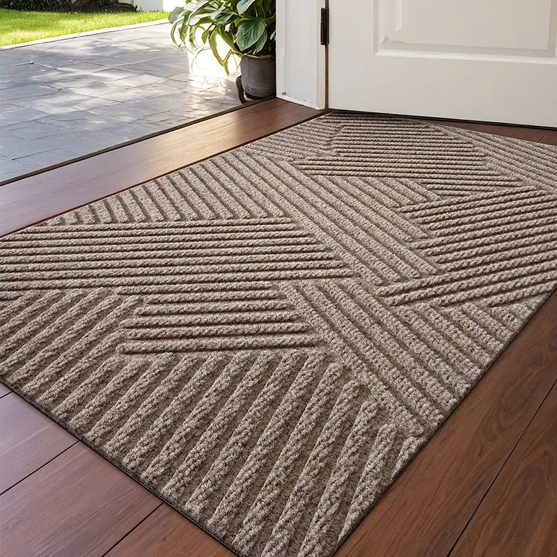 A rectangular gray textured doormat with a geometric pattern placed on a wooden floor near a white door.