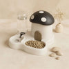 A white mushroom-shaped feeder and water dispenser with a black spotted cap, next to a glass of water and some pebbles.