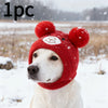 A white dog wearing a red knitted winter hat with bear ears in a snowy outdoor setting.