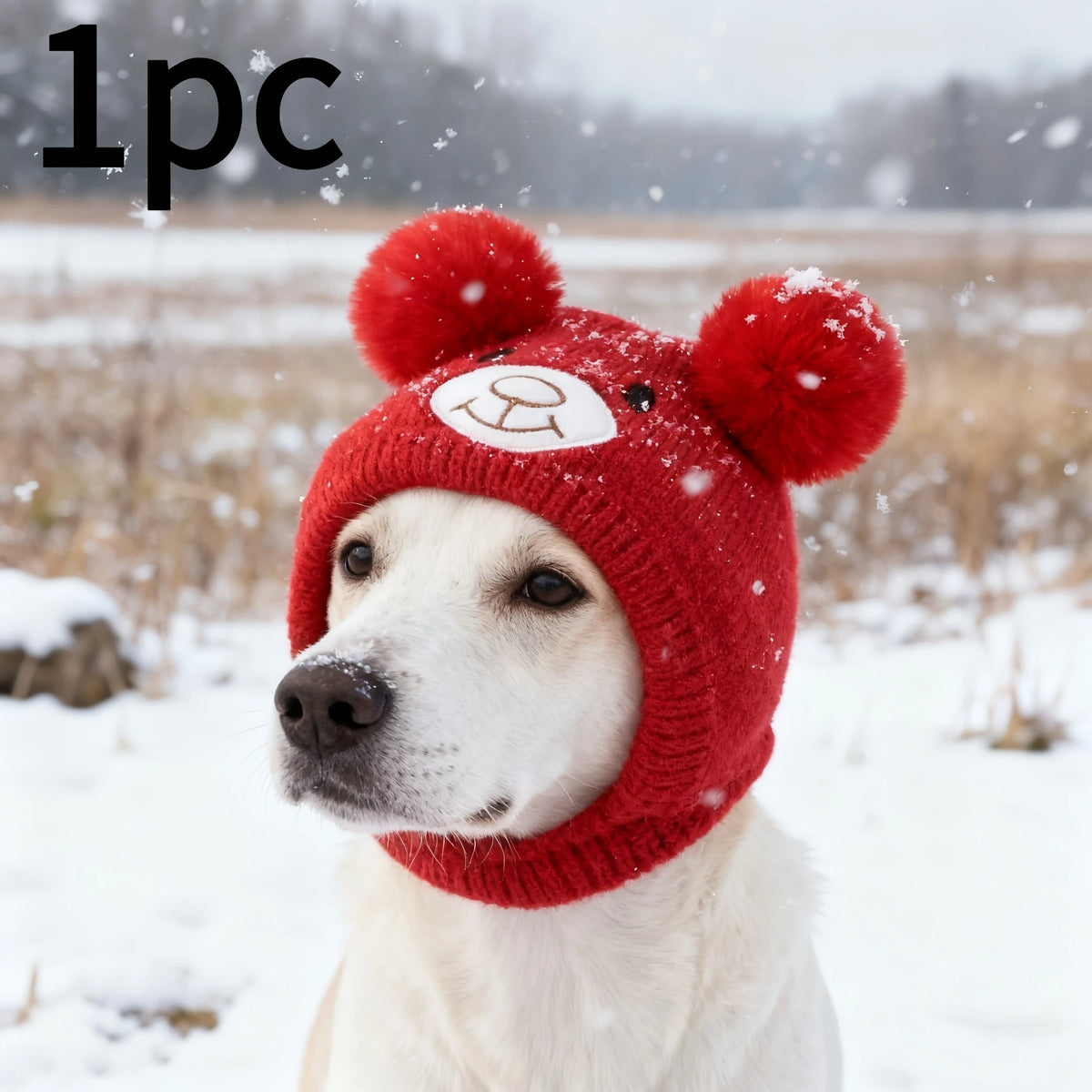 A white dog wearing a red knitted winter hat with bear ears in a snowy outdoor setting.